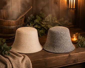May include: Two felt sauna hats, one beige and one grey, are displayed on a wooden surface. The hats have a classic bucket hat shape with a slightly flared brim. The background includes a wooden sauna interior, a wooden bucket, and greenery.