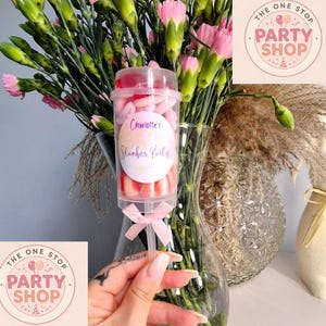 May include: A clear push pop filled with pink and white sweets, labelled "Charlotte's Slumber Party." The pop is held in front of a bouquet of pink carnations and greenery, with a "Party Shop" logo in the background.