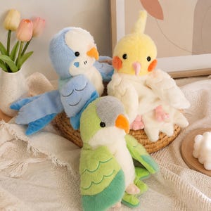 May include: Three plush bird toys featuring a blue and white parakeet, a yellow and white cockatiel with orange cheek patches, and a green and white parrot, each with black eyes, small beaks, and soft textured fabric wings and bodies.