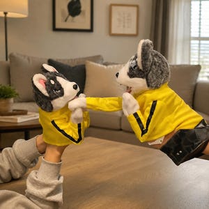 Puede incluir: Dos marionetas de peluche con forma de perro de pelaje gris y blanco, cada una con una chaqueta amarilla brillante con rayas negras en las mangas. La marioneta más grande lleva una falda negra, ambas tienen orejas rosadas y narices oscuras.