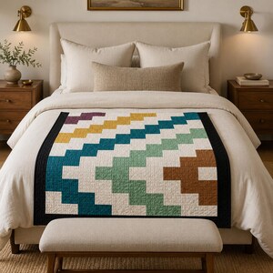 May include: A rectangular quilt featuring a geometric stair-step pattern in shades of teal, light green, mustard yellow, brown, and cream. The quilt is bordered by a solid black frame and is draped over a bed with cream-colored bedding.