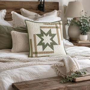 May include: A square throw pillow featuring a quilted cream-colored fabric with a central eight-pointed star design in olive green and tan. The surface displays intricate swirling stitch patterns throughout the fabric, finished with a green border.