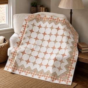 May include: A quilted blanket featuring a repeating pattern of white circular shapes and orange star-like accents. The border consists of a checkered design in orange and beige tones, with a zigzag edge that frames the central geometric motif.