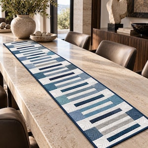 May include: A long rectangular table runner featuring a geometric pattern of alternating blue, navy, and white fabric strips. The design includes quilted sections with diamond textures and solid color blocks arranged in a repeating linear sequence.