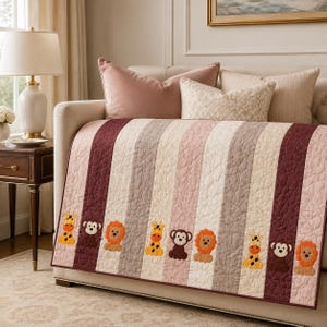 May include: A quilted throw blanket featuring vertical stripes in shades of burgundy, light pink, and cream. The blanket is decorated with repeating embroidered appliques of giraffes, monkeys, and lions positioned along the lower section.
