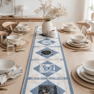 May include: A rectangular fabric table runner featuring a quilted pattern with blue and cream geometric borders. The design includes central square panels displaying a blue butterfly, floral motifs, and text, laid out on a light wood dining table.