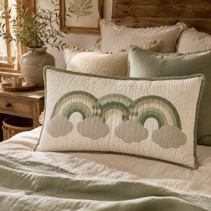 May include: A rectangular lumbar cushion featuring a quilted cream-coloured fabric with a repeating pattern of three sage green rainbows and grey clouds. The cushion has a sage green piped border and a soft, textured finish across the entire surface.