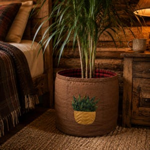 Könnte beinhalten: Ein zylindrischer Pflanzenkorb aus braunem Stoff mit einer aufgenähten Applikation einer grünen Pflanze in einem gelben Topf auf der Vorderseite. Das Innere des Korbes ist mit einem rot-schwarz karierten Stoff ausgekleidet und eine hohe grüne Pflanze steht darin.