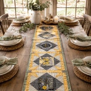 May include: A quilted table runner featuring a repeating diamond pattern in shades of yellow, gray, and white, decorated with printed butterfly and botanical motifs, laid across a rustic wooden dining table with place settings and greenery.
