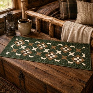 May include: A rectangular fabric table runner featuring a repeating geometric quilt pattern in shades of forest green, tan, and cream. The design consists of interconnected star shapes and small checkered squares, finished with a thin green border.