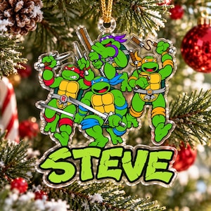 May include: A clear acrylic Christmas ornament featuring the four Teenage Mutant Ninja Turtles in green, yellow, and brown with their signature coloured masks and weapons, with the name STEVE printed in large green letters at the bottom.