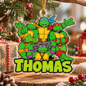 Pode incluir: Um enfeite de Natal em acrílico transparente com as quatro Tartarugas Ninja em verde, amarelo e castanho com as suas máscaras coloridas. O nome THOMAS está impresso em letras grandes de cor verde brilhante.