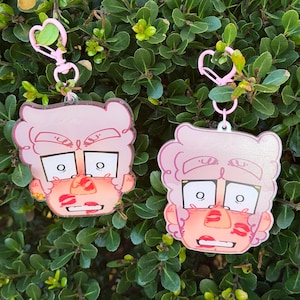 May include: Two pink acrylic keychains with a cartoon face design. The face has a light pink complexion, white-rimmed eyes, and a mouth covered in red lipstick kisses. Each keychain has a pink heart-shaped clasp.