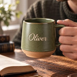May include: A sage green ceramic mug with a speckled finish and a natural, unglazed terracotta base. The name Oliver is engraved in a cursive script on the front of the mug. The mug features a sturdy handle and a smooth, rounded rim.