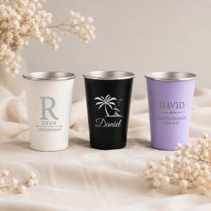 Könnte beinhalten: Drei Edelstahlbecher in Weiß, Schwarz und Helllila. Der weiße Becher zeigt den Buchstaben R, Ryan und Groomsman. Der schwarze Becher zeigt eine Palme und Daniel. Der lila Becher zeigt David, Groomsman und das Datum 15.08.2026.