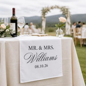 May include: A white table runner draped over a beige tablecloth, featuring the black printed text MR. & MRS. Williams and the date 08.10.26. The runner is positioned on a table with a wine bottle, two glasses, and a single peach rose in a vase.