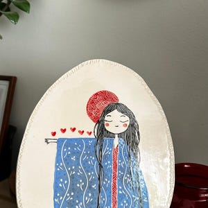 May include: Hand-painted ceramic plate with a woman in a blue kimono, red accents, and a red sun. The plate has a white background and a decorative edge. Another plate with a koi fish design is visible.