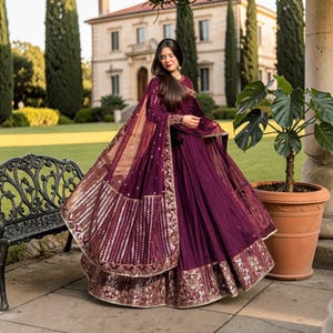 May include: A deep plum-colored floor-length gown featuring a full, pleated skirt and a matching sheer dupatta. The garment is adorned with intricate gold embroidery and sequin detailing along the wide hemline and the edges of the draped shawl.