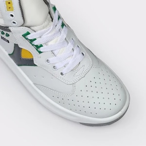 May include: A high-top sneaker featuring a white leather upper with a perforated toe box, white laces, and a thick white sole. The design includes accents in yellow, green, and dark gray, with a small yellow square detail near the top eyelets.