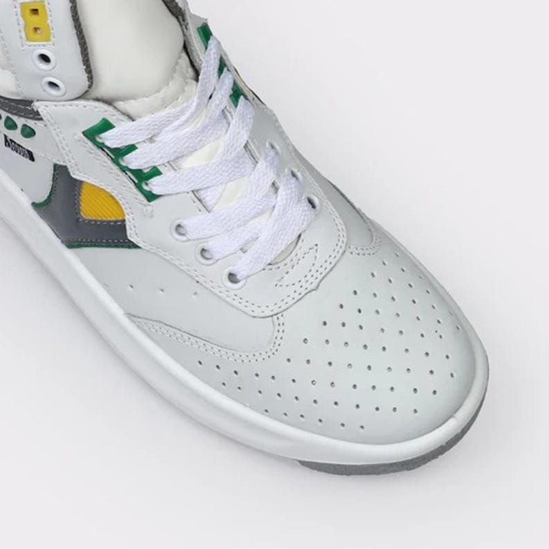 May include: A high-top sneaker featuring a white leather upper with a perforated toe box, white laces, and a thick white sole. The design includes accents in yellow, green, and dark gray, with a small yellow square detail near the top eyelets.