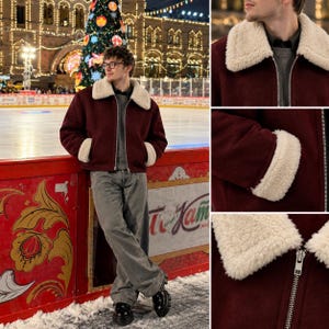 May include: A burgundy jacket featuring a white sherpa collar and matching sherpa cuffs on the sleeves. The jacket has a full-length silver metal zipper closure down the front and is worn over a grey sweater with grey trousers and black shoes.