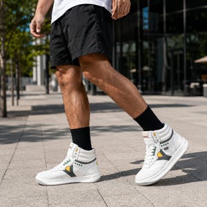 May include: High-top sneakers in white leather featuring perforated toe boxes, white laces, and dark gray side panels with yellow accents. The sneakers have a padded collar, a thick white rubber sole, and black socks worn underneath.