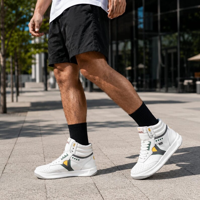 May include: High-top sneakers in white leather featuring perforated toe boxes, white laces, and dark gray side panels with yellow accents. The sneakers have a padded collar, a thick white rubber sole, and black socks worn underneath.