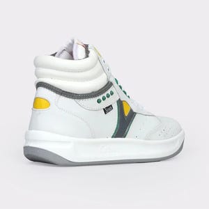 May include: A high-top sneaker in white leather featuring a padded collar, a thick white midsole, and a grey outsole. The shoe includes green eyelet accents, a yellow heel patch, and a dark grey chevron-style side panel with the text Sport.