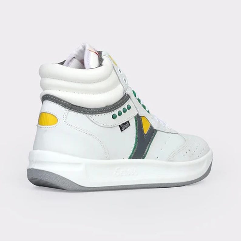 May include: A high-top sneaker in white leather featuring a padded collar, a thick white midsole, and a grey outsole. The shoe includes green eyelet accents, a yellow heel patch, and a dark grey chevron-style side panel with the text Sport.