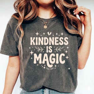 May include: A charcoal grey short-sleeve t-shirt featuring the cream-coloured text "KINDNESS IS MAGIC" printed on the front. The design includes decorative star and leaf accents surrounding the bold, rounded lettering in a centred layout.