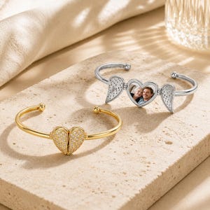 Op de afbeelding: Twee armbanden met hartvormige middenstukken en engelenvleugelontwerpen. De ene armband is goudkleurig met heldere steentjes, terwijl de andere zilverkleurig is en een kleine ronde foto van twee personen in het hartje laat zien.