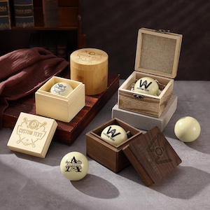 May include: Several off-white billiard balls featuring custom black text and graphics, including the word Anderson, displayed alongside various square and cylindrical wooden boxes with hinged lids, some engraved with the text CUSTOM TEXT.