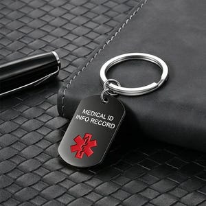 May include: A black metal dog tag keychain featuring a silver split ring. The tag displays the white text MEDICAL ID INFO RECORD above a red Star of Life medical symbol. The surface of the tag is smooth and rectangular with rounded corners.