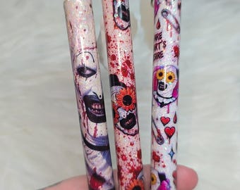 Könnte beinhalten: Drei einziehbare Stifte mit weißem Glitzer-Finish. Der erste Stift zeigt ein Clown-Gesicht mit roten Spritzern. Der zweite Stift hat rote Spritzer und orangefarbene Blumen. Der dritte Stift zeigt Herzen, Sicherheitsnadeln und den Text YOU ARE MY ART'S DESIRE.