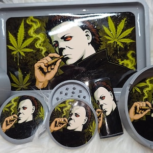 May include: A gray rolling tray, a circular grinder, a cylindrical container, and a round ashtray, all featuring a graphic illustration of a character with a white mask and dark hair smoking a cigar, surrounded by yellow-green cannabis leaf patterns.