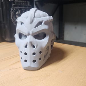 May include: A light gray, 3D-printed hockey-style mask featuring a skull-like design with prominent eye sockets, a central nose ridge, and a series of circular ventilation holes across the lower section, showcasing a matte, textured finish.