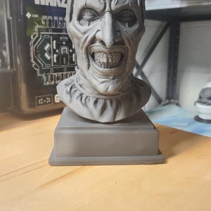 May include: A monochromatic gray 3D printed bust of a clown character featuring a tall hat, a ruffled collar, and a wide, toothy grin. The sculpture is mounted on a square base and displays detailed facial wrinkles and deep-set eyes.