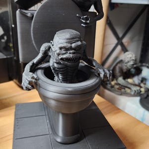 May include: A black 3D printed figurine featuring two creatures emerging from a toilet. One creature is inside the bowl with sharp teeth, while the other is perched on the tank. The base is rectangular with the word GHOULIES printed in red and white.