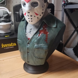 May include: A sculpted bust of a figure wearing a white hockey mask with red triangular markings and a dark green collared shirt. The shirt features a red blood-like stain on the chest area. The figure is mounted on a circular black base.