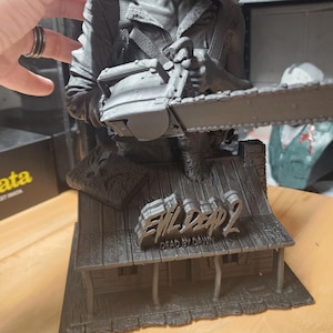 May include: A gray 3D-printed resin statue depicting a character holding a chainsaw, positioned above a cabin structure. The cabin roof features the text Evil Dead 2 Dead by Dawn and a book with a face design. The figure has a detailed facial expression.