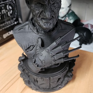 May include: A matte black bust of a scarred character wearing a fedora and a sweater with distressed details. The figure has a glove with four long, sharp metal-like blades extending from the fingers, resting on a circular base with wood-like texture.