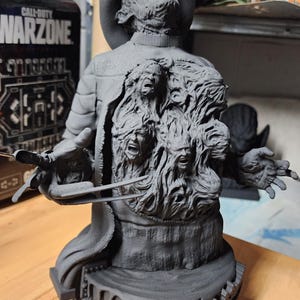 May include: A matte gray 3D printed bust of a character wearing a fedora and a sweater, featuring a chest area sculpted with multiple screaming faces and a right hand equipped with long, sharp metal-like blades extending from the fingers.