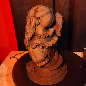 May include: A monochromatic brown bust sculpture of a figure with long hair tied in a ponytail, wearing a garment with a ruffled collar. The figure is positioned on a circular, tiered base that features a series of small, evenly spaced circular indentations.