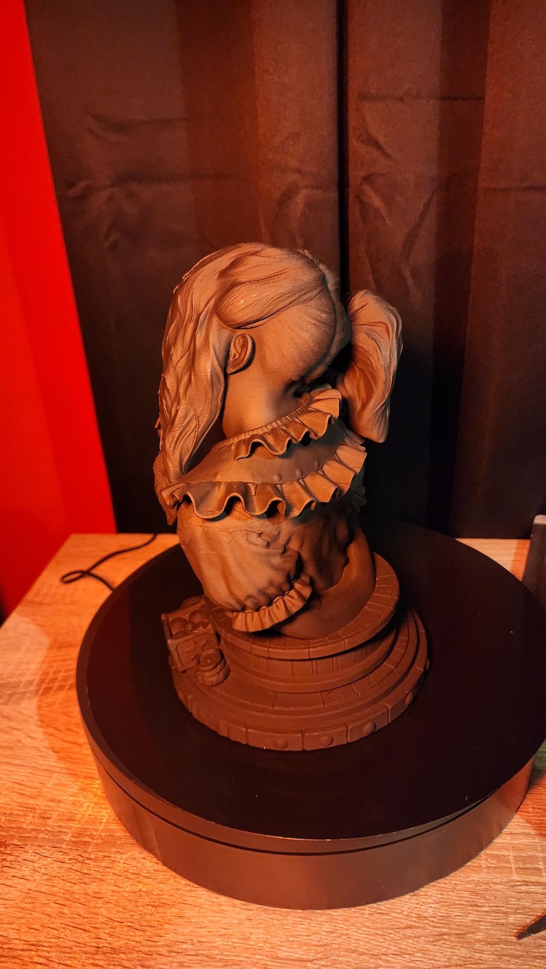 May include: A monochromatic brown bust sculpture of a figure with long hair tied in a ponytail, wearing a garment with a ruffled collar. The figure is positioned on a circular, tiered base that features a series of small, evenly spaced circular indentations.