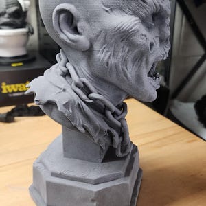May include: A grey 3D printed bust of a creature with a bald head, textured skin, and an open mouth showing teeth. The figure wears a sculpted chain around its neck and rests on a hexagonal base. The surface shows visible layer lines from printing.