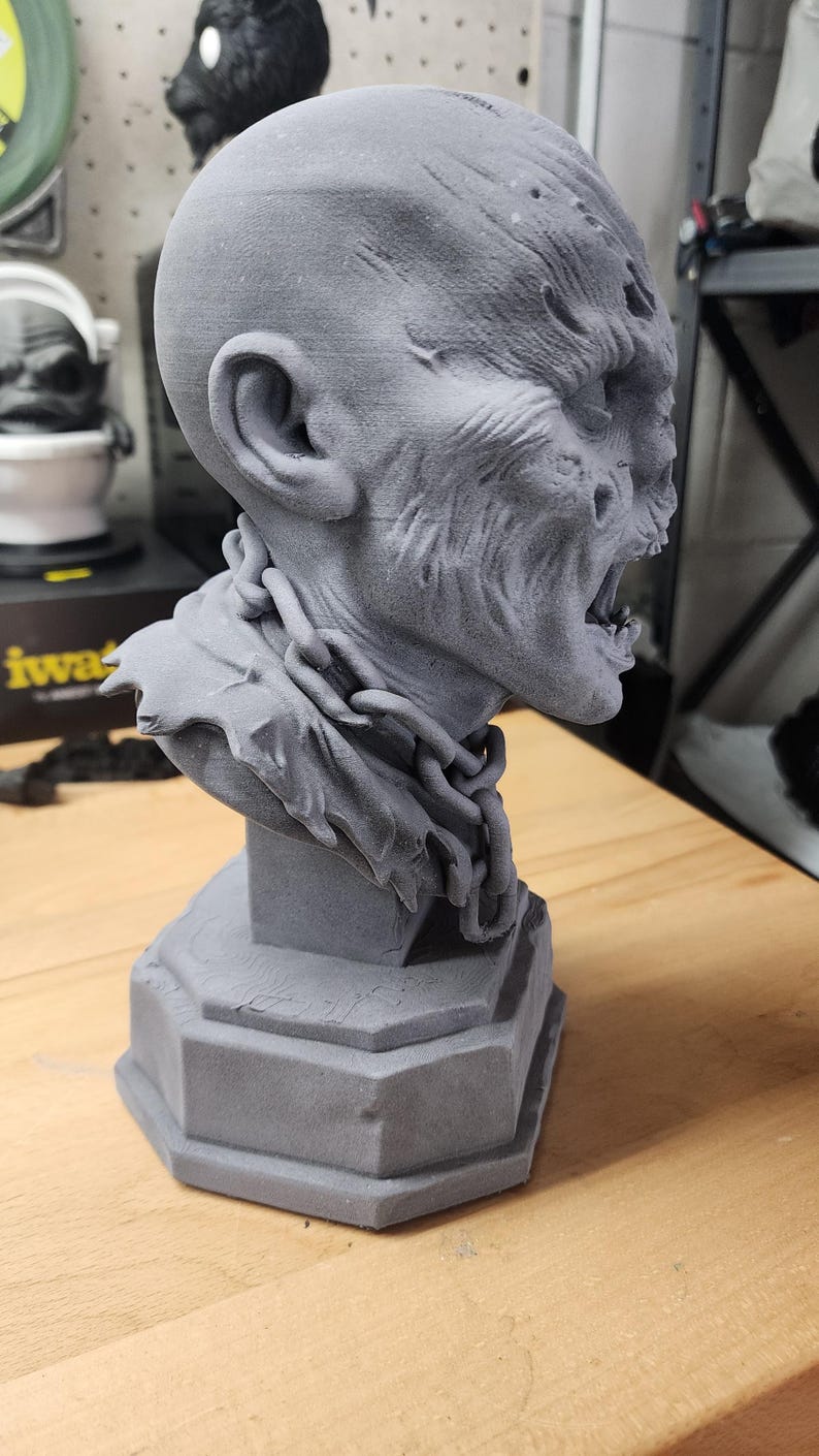 May include: A grey 3D printed bust of a creature with a bald head, textured skin, and an open mouth showing teeth. The figure wears a sculpted chain around its neck and rests on a hexagonal base. The surface shows visible layer lines from printing.