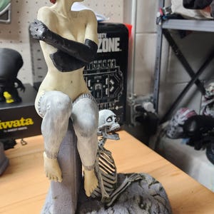 Puede incluir: Una figura pintada que muestra a un personaje con cabello corto rojo, guantes negros y medias blancas, sentado sobre un bloque gris que imita piedra. Un peque&ntilde;o esqueleto blanco se sit&uacute;a en la base del bloque sobre un suelo gris texturizado.