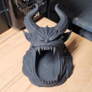 May include: A matte black 3D printed sculpture of a horned demon head with a wide, open mouth revealing sharp teeth. The creature features pointed ears, furrowed brows, and curved horns extending upward from the top of the head.