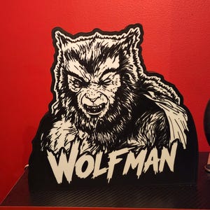 Peut inclure: Une enseigne en noir et blanc repr&eacute;sentant une illustration d&eacute;taill&eacute;e d'un loup-garou avec de la fourrure et des dents pointues. Le mot WOLFMAN est imprim&eacute; en grandes lettres capitales blanches en bas de l'enseigne, bord&eacute;e de noir.
