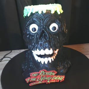 May include: A black, textured zombie head sculpture with bright green hair, wide white eyes, and white teeth. A red and green plaque at the base features the text "THE RETURN OF THE LIVING DEAD" in a stylized font. The figure has a glossy finish.